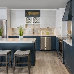 Kitchen Views - Kitchen showroom display featuring navy blue lower cabinetry, light gray upper cabinets with glass and X-pattern wine storage...