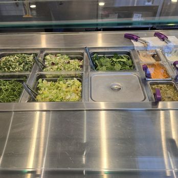 CAVA - A salad bar setup with stainless steel containers holding fresh leafy greens, lettuce, spinach, and other salad toppings. Tongs are placed in some containers for serving. The display is behind a glass shield, and the surface is made of stainless steel, typical of a cafeteria or restaurant salad station. - Mediterranean Near Me - Gambrills, Maryland