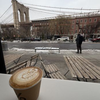 % Arabica - A latte with foam art is on a white surface inside a cafe, looking out onto a snowy street with parked cars, a person walking, and the Brooklyn Bridge in the background. - Coffee & Tea Near Me - Brooklyn, New York