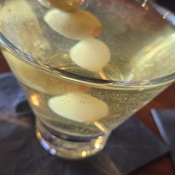 Murphy's Restaurant - Dirty martini - A clear cocktail served in a martini glass, garnished with green olives on a skewer, placed on a dark napkin on a wooden table. - Seafood Near Me - Prescott, Arizona