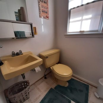 Benperlas - Bathroom Before - The bathroom contains a yellow toilet and a matching yellow sink with a mirror cabinet above it. A small basket is placed under the sink. There is a window with blinds allowing natural light to enter, casting shadows on the wall. The floor has two dark-colored mats, and a poster is hung on the wall near the mirror cabinet. - Handyman Near Me - Spencerville, Maryland