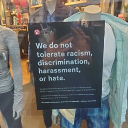 A sign is displayed in a retail store window, positioned among mannequins dressed in athletic apparel. The sign features a message condemning racism, discrimination, harassment, and hate, emphasizing values of respect, inclusion, and empathy. The store interior with clothing displays and mannequins is visible through the glass, and the sign is prominently placed to communicate the store's social stance.
