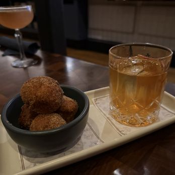 The Henry - Doughnuts & whiskey from holiday menu - Fried food in a black bowl and a whiskey cocktail with ice on a tray are placed on a wooden table in a restaurant or bar. - New American Near Me - Phoenix, Arizona