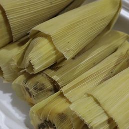 Green Corn Tamales - Multiple green corn tamales wrapped in corn husks are arranged on a white plate. The tamales are prepared with corn husks, and some filling is visible at the ends. The image focuses on the tamales, which are a traditional Mexican dish, and the setting appears to be a kitchen or dining area.