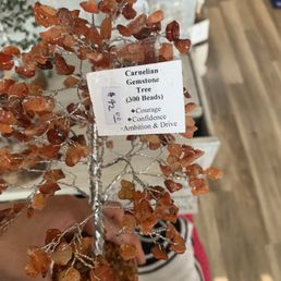 A decorative gemstone tree crafted from orange Carnelian stones, with a wire trunk and branches. The tree is mounted on a wooden base and has a label indicating it contains 300 beads, emphasizing qualities like courage, confidence, ambition, and drive. The background suggests an indoor retail environment with other items and wooden flooring.