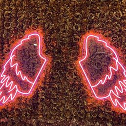 Background - The image features two neon pink wing outlines mounted on a wall densely covered with dark roses. The neon wings are symmetrical and outlined with bright pink light, contrasting with the dark, textured floral background. The roses are tightly packed, providing a rich, textured surface that enhances the visual impact of the neon light. The overall scene combines floral decor with illuminated art, suitable for photo opportunities or decorative purposes.