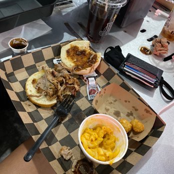 BP Smokehouse - Pulled pork sandwich Mac and cheese and tater tots - A food tray contains pulled pork sandwiches on buns, macaroni and cheese, tater tots, and various sauces, with drinks and condiments on a table. - Barbeque Near Me - Tomah, Wisconsin
