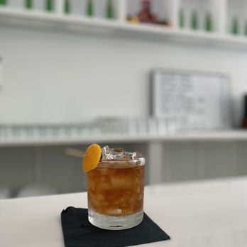 Imaginary Factory - Boozy Old Fashioned - A glass of cocktail with ice and an orange slice garnish sits on a black napkin at a bar counter. - Cocktail Bars Near Me - Madison, Wisconsin
