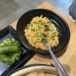 Dan Dan Noodles - A black bowl contains Dan Dan noodles topped with chopped green onions, placed on a wooden table. A side dish of sliced cucumbers is nearby.