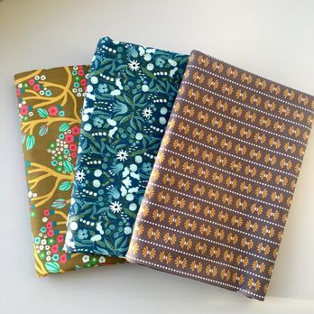 Harmony - Three fabric pieces are arranged on a white surface, each featuring different patterns. The first fabric has a brown background with pink, white, and green floral motifs. The second fabric displays a dark blue background with white and light blue floral and leaf patterns. The third fabric has a brown background with a repeating pattern of orange and white geometric shapes. The fabrics are neatly laid out, highlighting their colorful and varied designs. - Fabric Stores Near Me - Provo, Utah