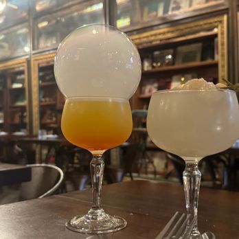 Tavern 157 - Two cocktails with unique garnishes are placed on a wooden table inside a bar with wooden shelves and framed pictures in the background. - Lounges Near Me - Flushing, New York