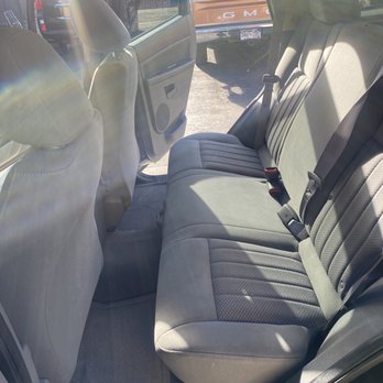 TruTouch - My dogs seat. - The image shows the interior of a vehicle focusing on the backseat area, which has gray fabric upholstery with a textured pattern on the seat cushions. Seat belts are visible, including a three-point belt on the right side. The door panel on the right side features a handle and a speaker grille. Sunlight streams into the vehicle, creating lens flare effects. Outside the vehicle, other parked cars and a tree are visible, indicating an outdoor parking area. - Auto Detailing Near Me - Denver, Colorado