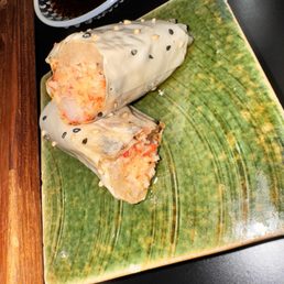 Crab dynamite - A sushi roll with a white sesame seed topping is shown on a green ceramic plate. The roll is partially eaten, revealing a filling that appears to include seafood and rice. The plate is on a black surface, with a small dish of soy sauce and other tableware visible in the background.