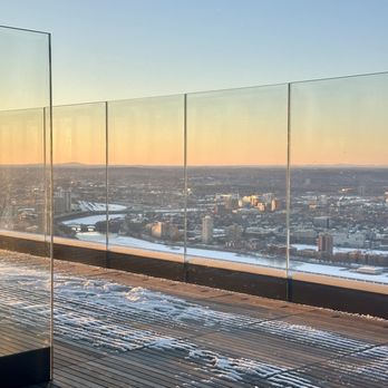 View Boston - Outdoor viewing platform - An outdoor rooftop viewing platform with glass panels offers a panoramic view of a city skyline during sunset, with snow on the wooden deck. - Observatories Near Me - Boston, Massachusetts