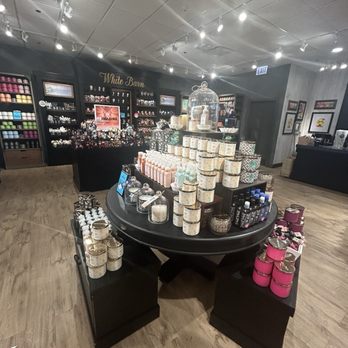 Bath & Body Works - The image shows a circular display table in a Bath & Body Works store, featuring various candles and bath products arranged in stacks. The background includes shelves filled with colorful candles and body care items. The store has a modern interior with a ceiling equipped with track lighting, and the floor is wood-like. The store's signage, 'White Barn,' is visible on the back wall, indicating the brand's section. - Cosmetics & Beauty Supply Near Me - Chicago, Illinois