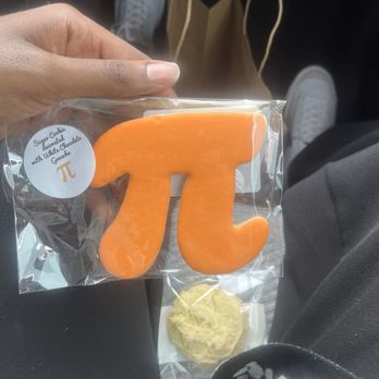 Bake Share Baking - pi day shortbread cookie ($3.14) - A person is seated, holding a clear plastic bag with an orange cookie shaped like the pi symbol, labeled as a sugar cookie decorated with white chocolate. The background shows a pair of white sneakers, a brown paper bag, and part of the person's clothing. The setting appears to be inside a vehicle. - Desserts Near Me - Grayslake, Illinois