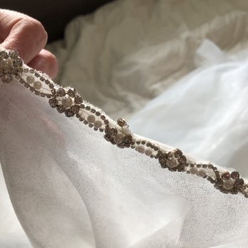 Custom Tailoring Services - The image shows a hand holding a piece of sheer, white fabric adorned with a decorative beaded trim featuring pearls and rhinestones. The background includes a soft, white, possibly satin or silk, fabric surface. The focus is on the intricate beadwork along the edge of the fabric, suggesting it may be part of a wedding dress or formal attire. - Sewing & Alterations Near Me - Brookfield, Wisconsin