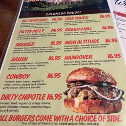 Large menu of burgers - The menu from Brush Creek Burgers displays a list of burger options including BC Avocado, Fire Truck, Patty Melt, Green Chili, and others, each with detailed descriptions and prices ranging from $14.95 to $16.95. It emphasizes that all burgers come with a choice of side, such as French fries, sweet potato fries, or coleslaw. The menu features a rustic design with a scenic background image at the top and a large burger photo on the right side, providing a clear view of the food offerings and pricing.
