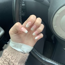 Ombré nails - The image shows a person's left hand with long, natural, well-maintained nails, positioned near the steering wheel of a car. The person is wearing a beige knitted sweater and a white shirt cuff. The interior of the vehicle includes a black steering wheel and dashboard controls, with part of the instrument panel visible.