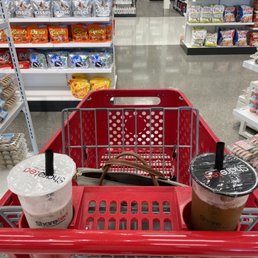 Nothing hits better than a boba while shopping at Target - A red shopping cart holds two iced beverages with black straws, positioned in a grocery store aisle. The background features shelves stocked with snack items such as Reese's, Hershey's, and other packaged foods, with a clean, well-lit store environment and visible product displays.