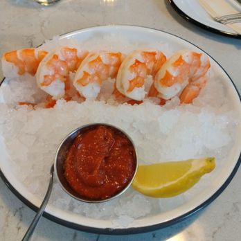 Out There - A round white plate contains cooked shrimp arranged on a bed of crushed ice, accompanied by a small metal cup of cocktail sauce and a lemon wedge. The plate is set on a marble table with a glass of water, a lit candle, and a set of silverware on a plate in the background. The setting appears to be a restaurant table. - Wine Bars Near Me - Sawyer, Michigan
