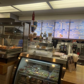 Charcoal Delights - Menu area, salad/pastry bar - Counter area in a casual restaurant with a glass display case containing pre-packaged salads and desserts, a menu board listing various food options, and a beverage station with coffee and drink dispensers. The background includes a ceiling with fluorescent lighting and a tiled wall behind the counter, with disposable cups and utensils available for customers. - Burgers Near Me - Des Plaines, Illinois