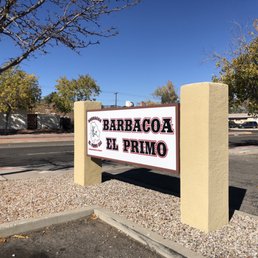 A rectangular sign for Barbacoa El Primo is mounted on two beige pillars beside a street in Albuquerque, NM, with trees and buildings in the background.