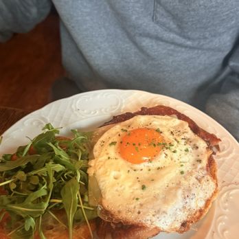 Longman & Eagle - Breakfast sandwich - A breakfast plate includes a fried egg with a runny yolk, seasoned with herbs, placed on crispy bacon strips. Next to the egg, there is a small serving of fresh greens, possibly arugula, on a white decorative plate with embossed patterns. The plate is held by a person wearing a gray zip-up sweater. - Gastropubs Near Me - Chicago, Illinois