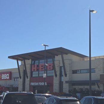 H-E-B Rockwall - Hello, dear friend! - A wide-angle view of the front of an H-E-B grocery store, featuring prominent store signage, a modern architectural facade with large windows, and a parking lot filled with vehicles. The building has a distinctive roof design with angled supports, and the sky is clear and blue, indicating a sunny day. - Grocery Near Me - Rockwall, Texas