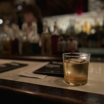 The Meadowlark - A glass of whiskey with ice sits on a marble bar counter, with a blurred background of bottles and bar equipment. - Speakeasies Near Me - Chicago, Illinois
