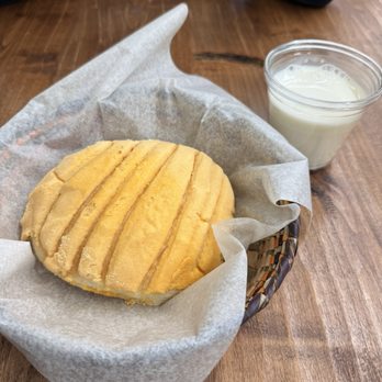 Cafe es Amor - Concha with Milk - A round, yellow concha pastry with ridges is placed in a basket lined with parchment paper, accompanied by a cup of milk on a wooden table. - Coffee & Tea Near Me - Albuquerque, New Mexico