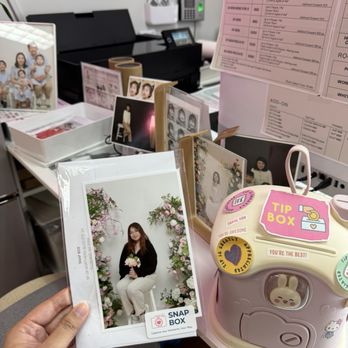 Snap Box - Complimentary Print - A person holds a printed photo of a woman sitting in front of a floral backdrop at a photo studio. The photo is in a clear plastic sleeve with branding and a QR code. The background includes a photo printing station with sample photos, a tip box with stickers, and a pink pricing guide. The setting appears to be an indoor photo studio or printing area. - Session Photography Near Me - Flushing, New York