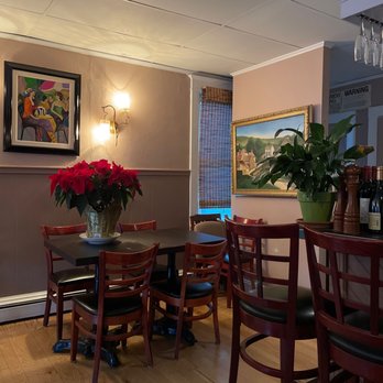 Cafe Les Baux - a table and chairs in a restaurant - The image shows a section of a restaurant interior featuring a wooden dining table with six chairs, some with black cushions. A large potted plant is placed on a counter, and framed artwork decorates the walls. The lighting includes wall-mounted fixtures, and a window with a bamboo shade is visible. The flooring is wooden, and the ceiling has a simple, tiled design. The setting suggests a casual dining environment with decorative elements. - French Near Me - Millbrook, New York