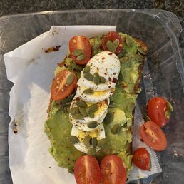 Avocado toast-so delicious! - A slice of avocado toast garnished with cherry tomato halves, sliced boiled eggs, pumpkin seeds, and red pepper flakes, served in a clear plastic container.