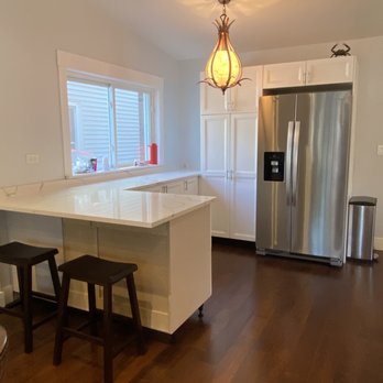 Pearlstone Cutters - The image shows a kitchen with white cabinets and a marble countertop island. A window above the counter allows natural light into the space, with a red object placed on the windowsill. A stainless steel refrigerator is positioned against the wall, with a small decorative item on top. The ceiling has a hanging light fixture that casts a warm glow, and the floor is made of dark wood. Two black stools are placed at the island, and a trash can is visible near the refrigerator. - Kitchen & Bath Near Me - Chicago, Illinois