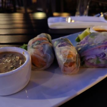 501 Local - Spring Rolls - The image shows a white rectangular plate with three spring rolls and two small bowls of dipping sauces, placed on a dark table. The background includes blurred elements of a restaurant environment, with some utensils and glasses visible. The lighting is dim, highlighting the food presentation. - New American Near Me - Winnetka, Illinois