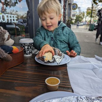 El Tiny Cafe - Feel the excitement as he dives into his burrito. - A young boy with blonde hair, wearing a teal jacket, is seated at an outdoor cafe table, holding a burrito wrapped in foil, with a cup of sauce nearby. - Cafes Near Me - Berkeley, California