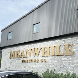 The photo depicts the front exterior of Meanwhile Brewing Co., with a brick wall displaying large gold signage reading 'MEANWHILE BREWING CO.' The building has a modern industrial design with gray metal siding and two small windows. In front, there are several cactus plants and part of a parked car is visible. The sky is partly cloudy, indicating daytime.