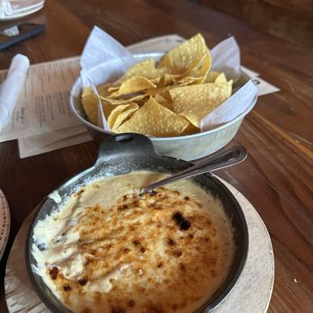 Bakersfield Mass Ave - Queso - A baked cheese dish with a golden-brown crust is served in a small skillet, accompanied by a bowl of tortilla chips on a wooden table. - Bars Near Me - Indianapolis, Indiana