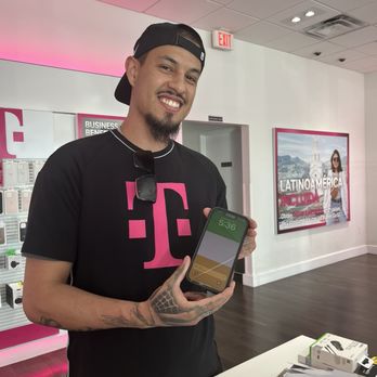 T-Mobile Experience Store - A man with tattoos, wearing a black cap backwards and a black T-Mobile shirt, displays a smartphone in a T-Mobile store. - Mobile Phones Near Me - Albuquerque, New Mexico