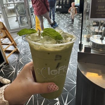 Lofty Coffee - Matcha latte - tasted of almonds and that's it - A hand with a polka-dotted nail holds a glass of iced matcha latte topped with mint leaves and ice cubes inside Lofty Coffee. The background shows a patterned floor, a few customers, and a child walking towards the seating area near the entrance, with large glass doors and windows allowing natural light. - Coffee & Tea Near Me - Carlsbad, California