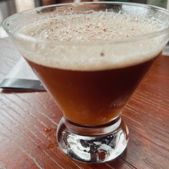 Station 1846 - A dark cocktail with a foamy top is presented in a glass on a wooden table inside a restaurant or bar setting. - Cocktail Bars Near Me - Milwaukee, Wisconsin