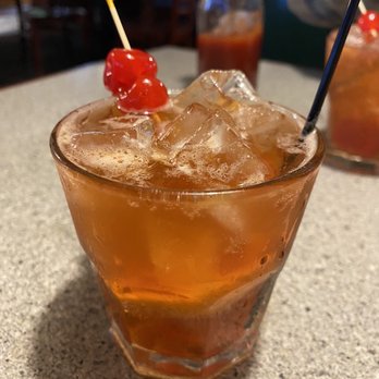 Cafe 20 - Old fashioned - A beverage in a short glass filled with ice cubes, topped with a cherry garnish on a cocktail pick. The drink has an amber color and is placed on a textured table surface. In the background, there are bottles and other drink containers, indicating a bar or restaurant setting. - American Near Me - Marengo, Illinois