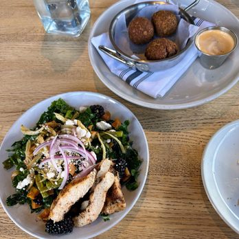 Showroom - Roasted vegetable salad (split into 2 plates, so this is 1/2) and chicken pot pie fritters - A white plate contains a mixed salad with greens, grilled chicken strips, sliced red onions, and blackberries. Next to it, a basket with three fried croquettes is placed on a white tray with a folded napkin, accompanied by a small cup of dipping sauce. A glass of water and part of another plate are visible on a wooden table surface. - New American Near Me - Frederick, Maryland