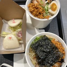 Spicy Tonkotsu - Two ramen bowls with toppings like seaweed, egg, and meat, accompanied by a small box of steamed buns with pink candles, on a tray.
