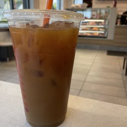 Iced coffee with oat milk - A plastic cup containing iced coffee with oat milk and ice cubes, with a straw inserted, is positioned on a light-colored table. The background shows the interior of a cafe, including a display case with baked goods and digital menu screens mounted on the wall. Natural light enters through windows, illuminating the scene.