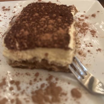 Sam's Kitchen - Tiramisu - A slice of tiramisu with layers of cream and coffee-soaked sponge, topped with cocoa powder, on a white plate. - Italian Near Me - Woburn, Massachusetts