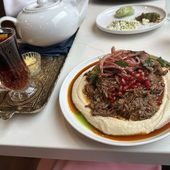 Joon - Joon: their tea is very good - A plate of Persian cuisine displays shredded beef garnished with pomegranate seeds and sliced onions, served on a bed of hummus with a sauce around the edge. A glass of tea with a lemon slice and a small dish of condiments are also visible on the table, indicating a dining experience at a Persian-themed restaurant. - Persian/Iranian Near Me - Vienna, Virginia