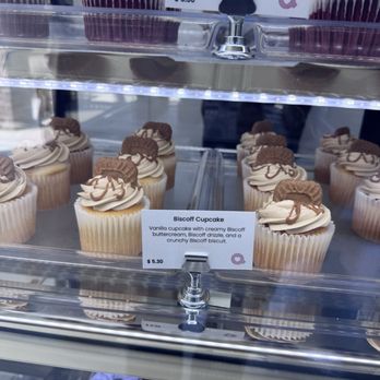 Arsty Cakery - Cupcakes with vanilla and Biscoff flavor are arranged in a display case, topped with cream and Biscoff cookies, with a label describing the flavor. - Bakeries Near Me - Coppell, Texas