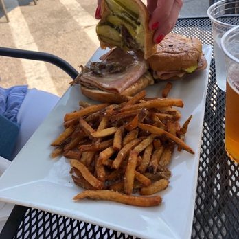 Commonwealth - Cubano - A rectangular white plate contains a serving of French fries, two sandwiches with different fillings, and sliced pickles. The plate is placed on a black metal outdoor table, with a person’s hand holding a sandwich above the plate. The background shows an outdoor seating area with chairs and a concrete surface, indicating an outdoor dining setting. - American Near Me - Chicago, Illinois